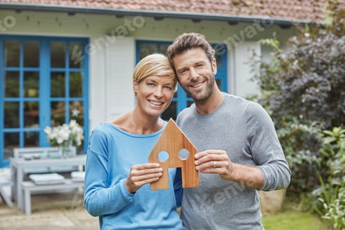 Preview: Portrait of smiling couple standing in front of their home holding house model