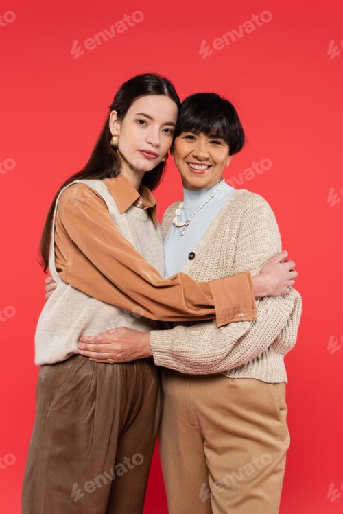 Preview: asian mother and daughter in stylish casual outfit embracing and looking at camera isolated on coral