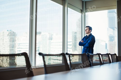 Preview: A man standing with arms crossed in a meeting room looking out of a window at an urban landscape.