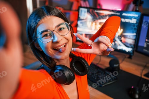 Preview: Smiling, making victory gesture and doing selfie. Young beautiful game streamer is indoors with pc