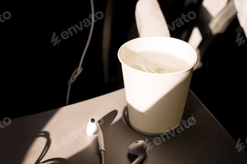 Preview: White paper cup of water and headphones on a folding table in an airplane. Drinks on the plane
