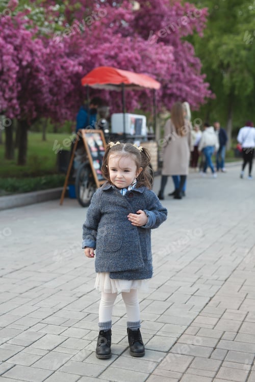 Preview: Little cute girl in a coat walks in the park against the background of a flowering tree in spring.