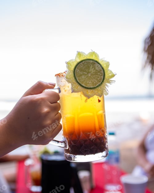 Preview: Female hand holding a refreshing and colorful Michelada drink garnished with lime.