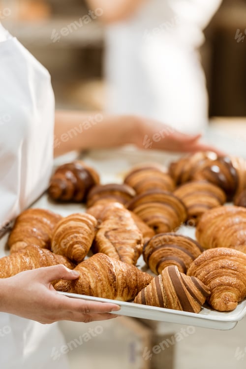 Preview: cropped shot of female baker holding tray with croissants on baking manufacture