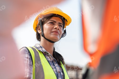 Preview: Confident female Indian engineer wearing protective helmet and vest working in construction site.