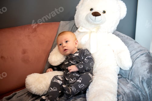 Preview: thoughtful cute newborn baby boy and big white teddy bear sitting on sofa or couch