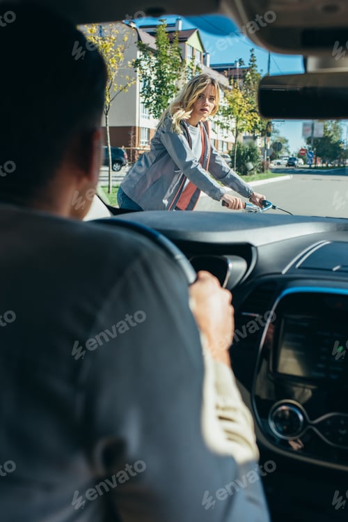 Preview: car driver waiting while young woman on bicycle crossing road