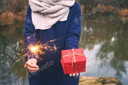 Preview: Woman holding sparkler and box gift in forest, winter day