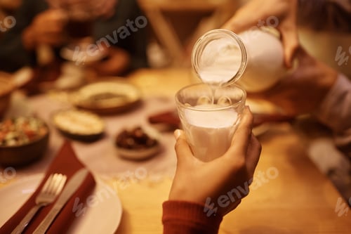 Preview: Close up of Muslim girl holding drinking glass while father is pouring her milk at dining table.
