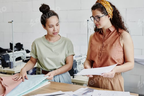 Preview: Young seamstress looking at fashion sketch on paper held by her colleague