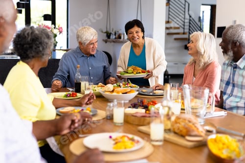 Preview: Smiling biracial senior woman serving food to multiracial friends at dining table in nursing home