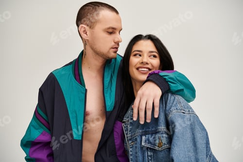 Preview: Young couple embraces with joy, showcasing their fashionable outfits in a studio backdrop.