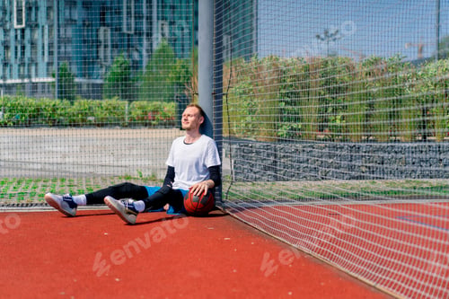 Preview: A tall guy basketball player with a ball in his hand sits leaning on the net of a basketball court