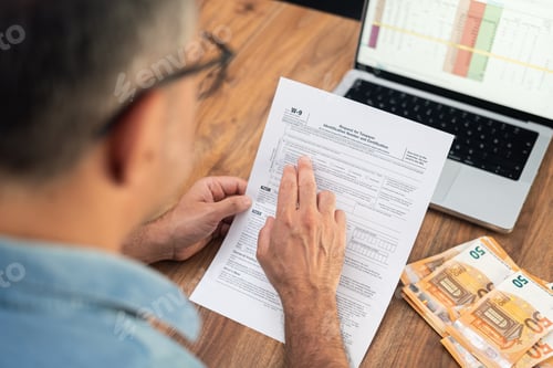 Preview: Small business owner examines W-9 tax form at table alongside laptop and euro bills