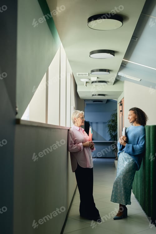 Preview: Mature Businesswoman Conversing with Younger Female Coworker in Office Center Hallway