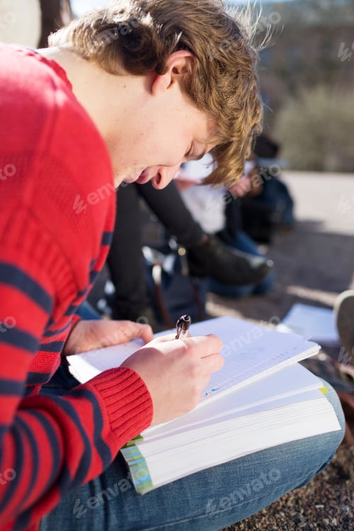 Preview: Teenage boy writing on book while sitting in college campus