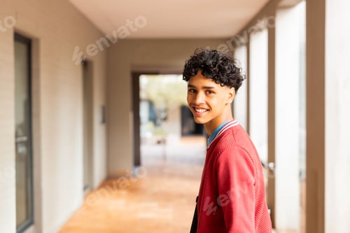 Preview: Teenage boy standing and looking back with smile wearing red jacket in campus corridor, copy space
