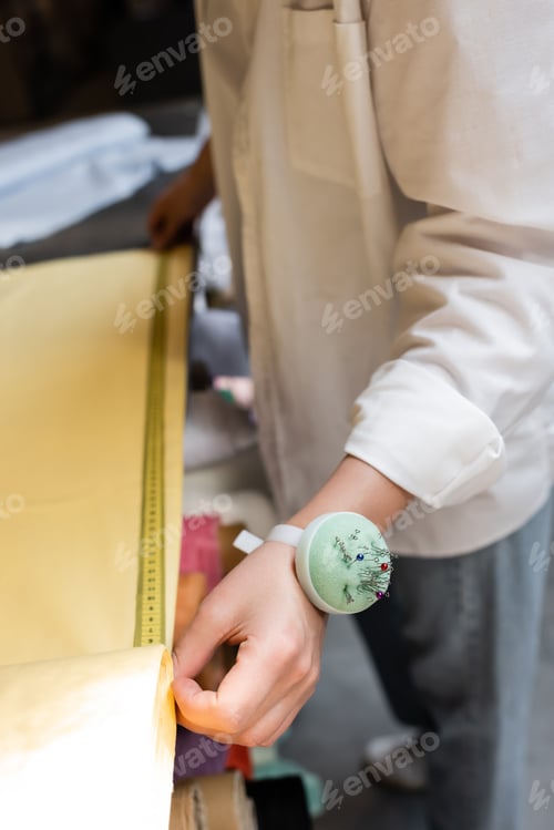 Preview: partial view of saleswoman with needle cushion on hand measuring yellow fabric in textile shop