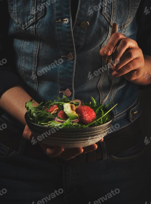 Preview: Healthy vegan lunch. Woman in blue denim holds ceramic bowl with avocado, arugula, grapefruit