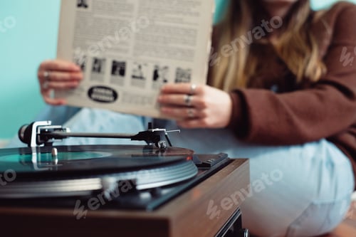 Preview: Young woman sitting in front of retro record player holding a record sleeve and listening to music