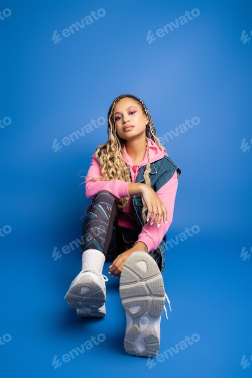 Preview: confident african american woman with dreadlocks sitting in sporty outfit on blue background