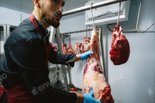 Preview: Male butcher standing by meat hanging on hook in meat shop
