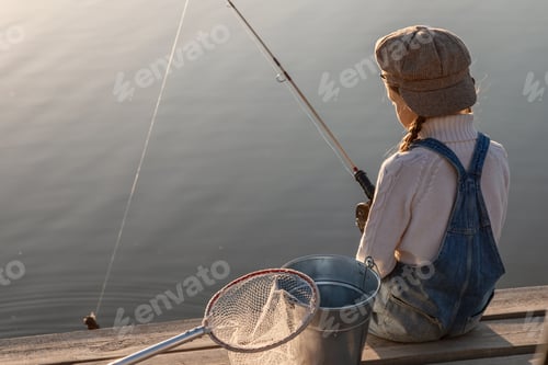 Preview: a girl in a cap with a bucket and a net is fishing on a lake with a wooden pier at sunset