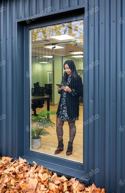 Preview: Hispanic woman working with a tablet behind of large window in office made from shipping containers