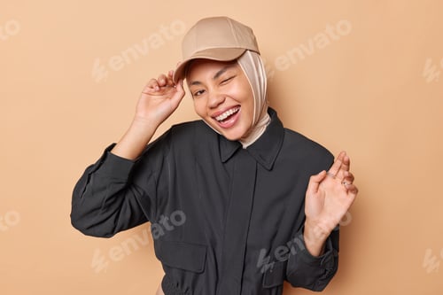 Preview: Happy positive woman wears cap on hijab black jacket smiles gladfully and winks eye stands glad