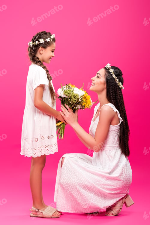 Preview: side view of adorable smiling daughter presenting beautiful flowers to happy young mother on pink
