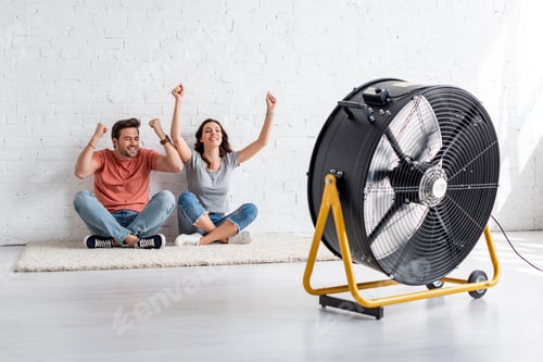 Preview: excited man and woman sitting on floor by white wall in front of blowing electric fan
