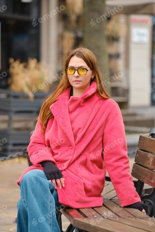 Preview: Young woman in pink coat and yellow sunglasses sitting on a bench in a city square during autumn
