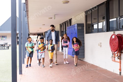 Preview: Diverse, happy male teacher and elementary schoolchildren walking in school corridor, copy space