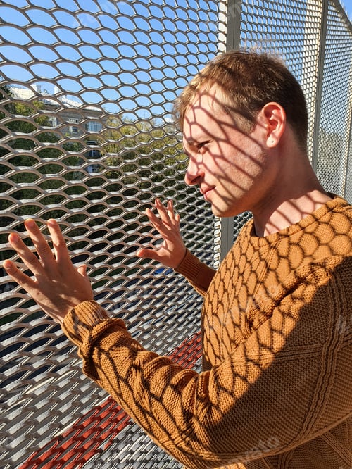 Preview: a young man stands behind a fence made of mesh. A shadow falls on the guy's face.