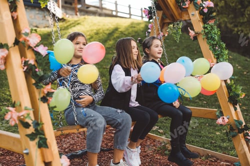 Preview: Three girls sitting on a swing by a lake, holding colorful balloons. The swing is decorated with flo