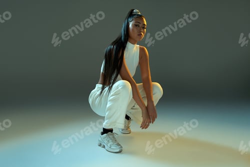 Preview: Full body of confident woman in white outfit with long dark ponytail hunkering down in studio