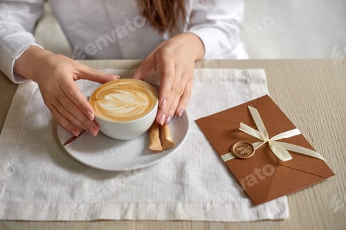 Preview: woman holding cup of cappuccino on light table in cafe and gift certificate in invitation envelope