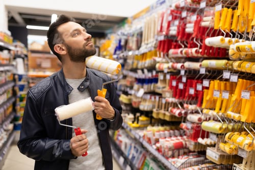 Preview: a man with different rollers for painting walls looks thoughtfully at a rack with brushes