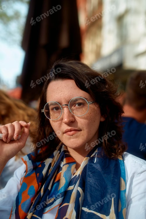 Preview: Confident woman with glasses and a colorful scarf seated outdoors in a cafe
