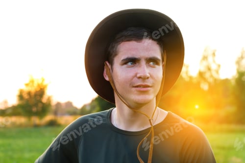Preview: Young man smiling farmer in cowboy hat at agricultural field on sunset with sun flare. Closeup portr