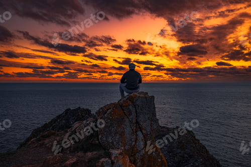 Preview: Rear of a male sitting on a pile of stones near seascape at sunset, dramatic sky background