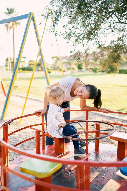 Preview: Mom leaned over to a little girl sitting on a carousel on the playground