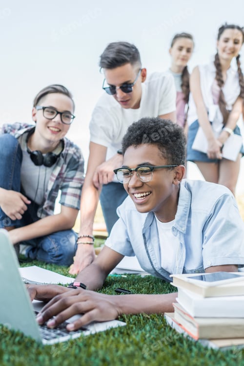 Preview: happy multiethnic teenage friends using laptop and studying in park
