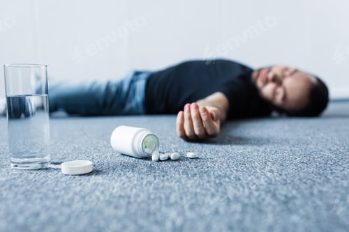 Preview: selective focus of unconscious man lying on grey floor near glass of water and container with pills