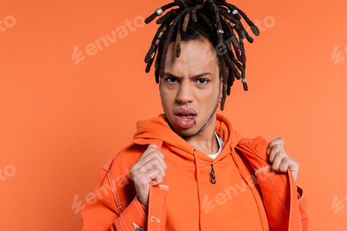Preview: displeased multiracial man with dreadlocks adjusting denim jacket isolated on coral background