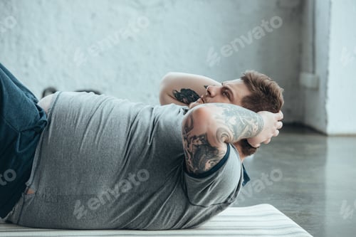 Preview: overweight tattooed man lying on fitness mat and doing sit up exercise at sports center