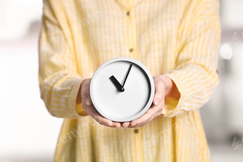 Preview: Young woman holding alarm clock on blurred background. Time concept