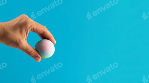 Preview: Woman holds colourful bath bomb carefully on blue background. Copy space