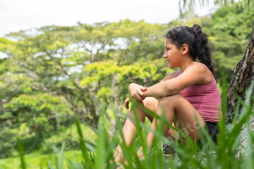Preview: latina brunette girl sitting next to a tree looking towards the horizon very pensive