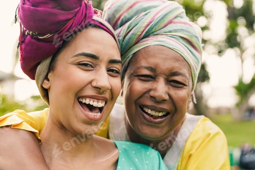 Preview: Happy african mother hugging adult daughter wearing traditional dress while smiling on camera